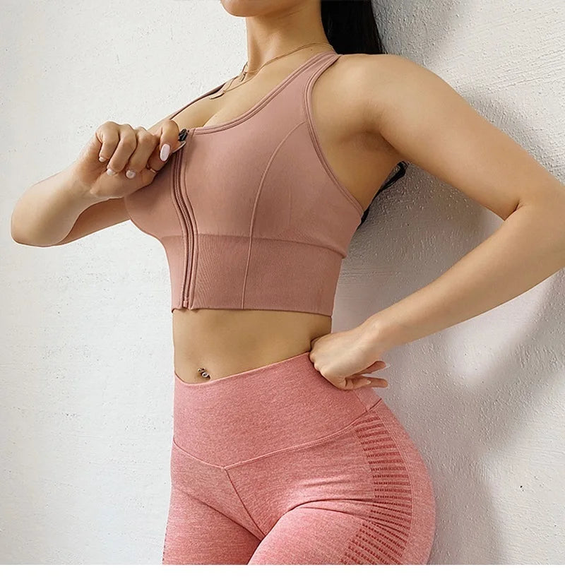 A woman wearing a dusty pink athletic sports bra and matching high-waisted leggings, posing against a light textured wall.