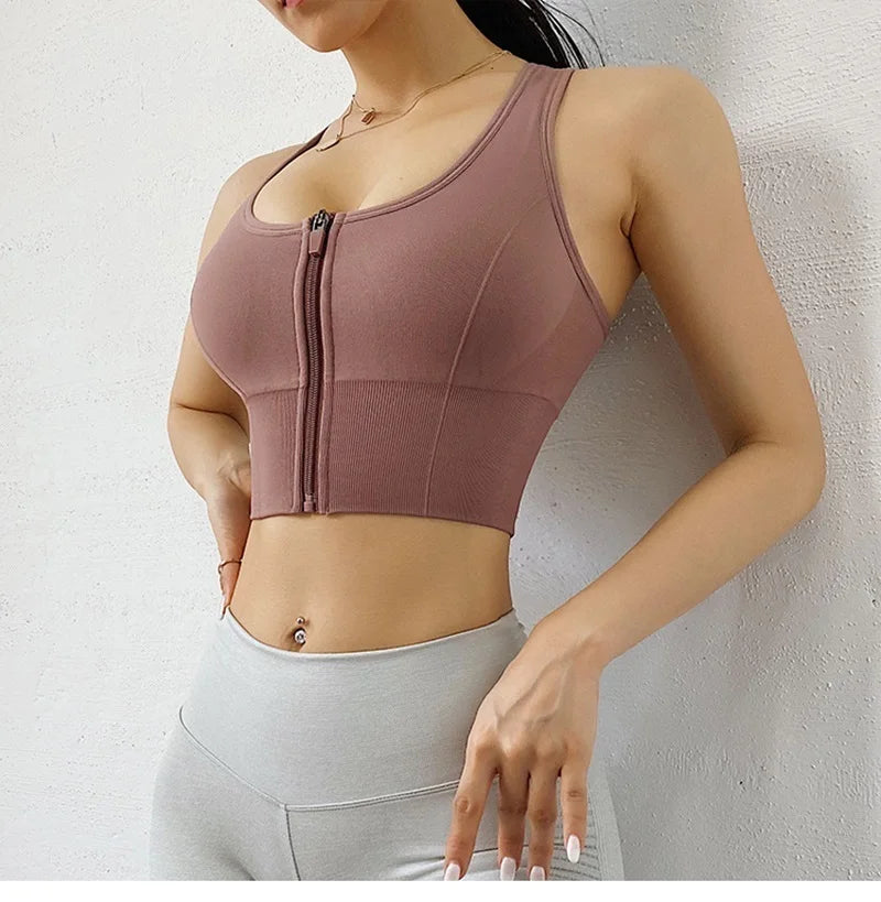 A woman wearing a sleeveless athletic crop top with a front zipper in a dusty mauve-brown color, paired with light gray leggings, posing against a textured white wall.