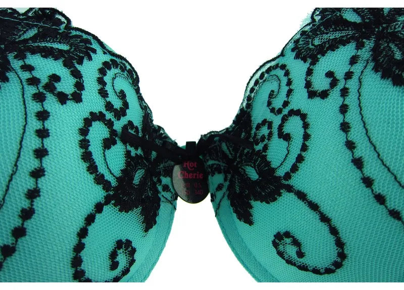 Close-up of a teal lace bra cup with black floral embroidery and a small central label, shown against a white background.