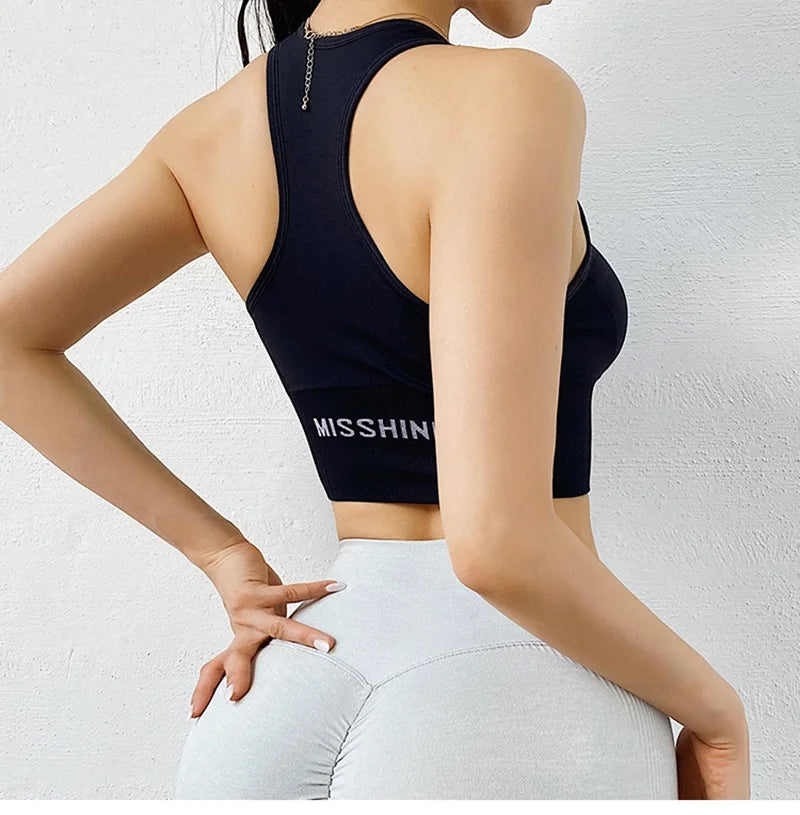 A woman shown from the back wearing a dark navy sports bra with 'MISSHIN' printed in white on the lower back, paired with light gray leggings, against a textured white wall.