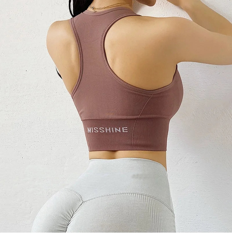 A woman shown from the back wearing a mauve-brown athletic sports bra with the brand name 'MISSHINE' printed in white on the lower back, paired with light gray leggings against a white textured wall.