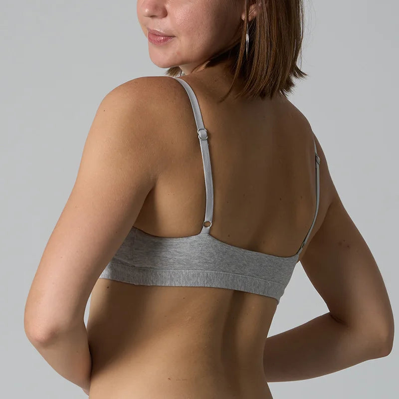A woman shown from the back wearing a light gray heather bralette with adjustable straps and a wide underband, against a plain light gray studio background.