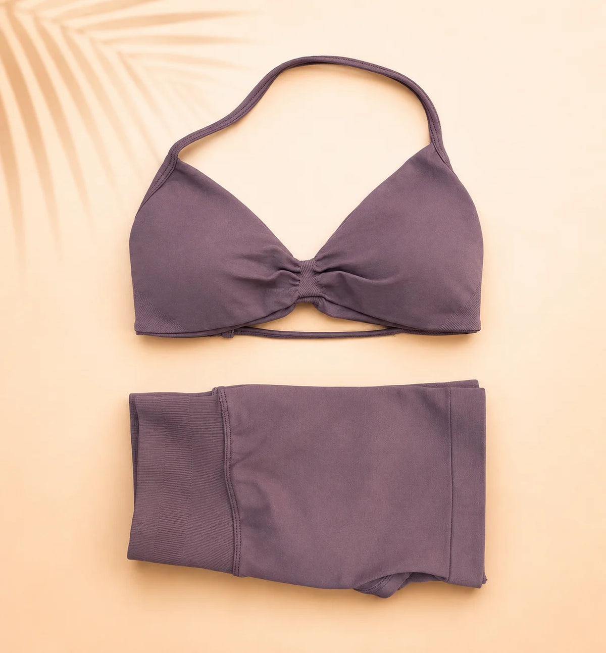 A two-piece bikini set in a muted plum purple color, featuring a halter-style triangle top with a front twist detail and matching high-waisted shorts, laid flat on a warm beige background with a palm leaf shadow.