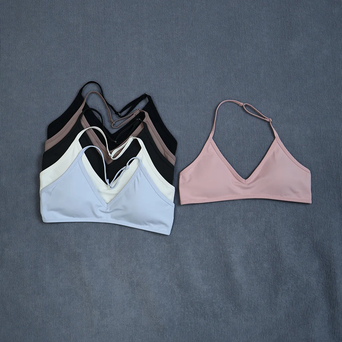 Four women's sports bra or bikini-style top pieces laid flat on a textured gray fabric background. The stack on the left shows multiple color options including black, taupe, white, and light blue, while a single light pink top is displayed to the right.