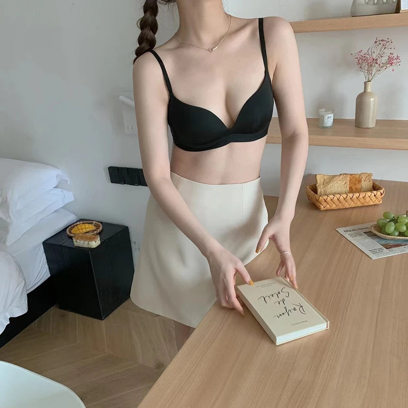 A woman wearing a black bra and a cream skirt stands at a wooden table in a bedroom setting, holding a book.