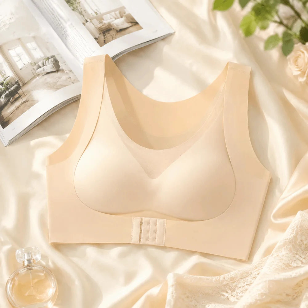 A beige wireless bralette with wide shoulder straps and a wide underband, styled on a bed of cream satin with an open magazine, a small perfume bottle, and a lace garment.