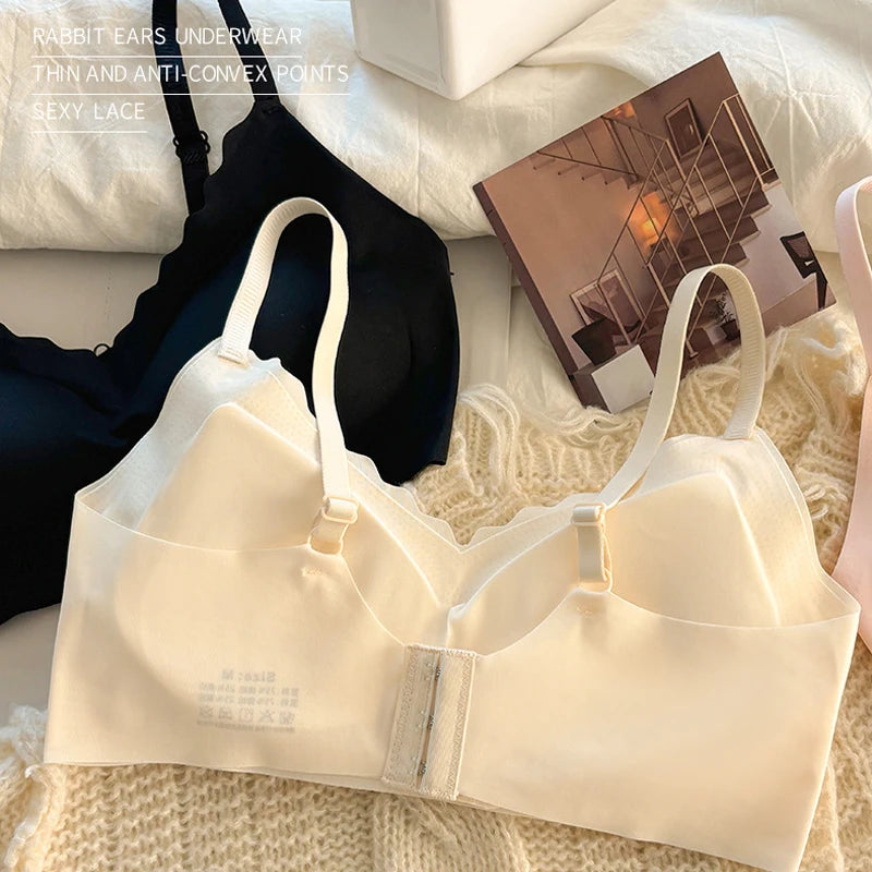 A cream-colored wireless underwire bra with adjustable straps and a center hook closure, styled on a textured beige knit blanket alongside a black bra and a magazine on a white background.
