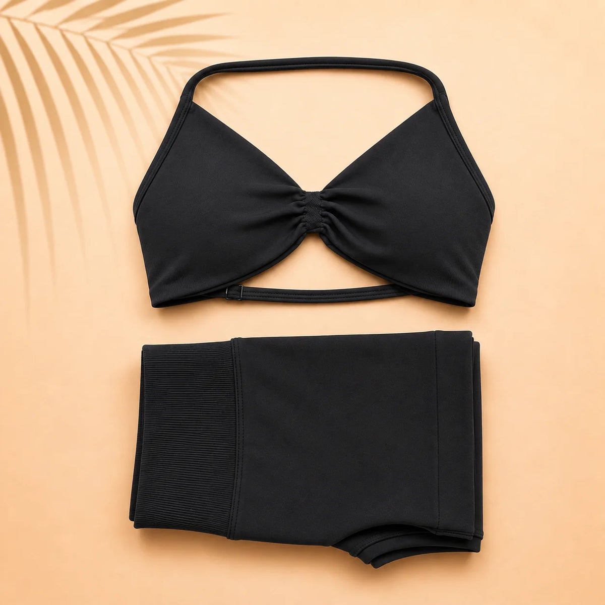 A black two-piece bikini set laid flat on a warm peach background. The top is a halter-style triangle bikini top with a gathered center front, and the bottom is a high-waisted ribbed bikini bottom.