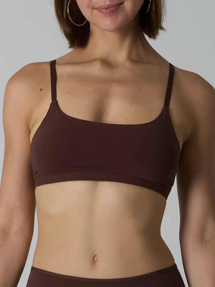 A close-up of a woman wearing a dark brown scoop-neck sports bra against a solid gray background.