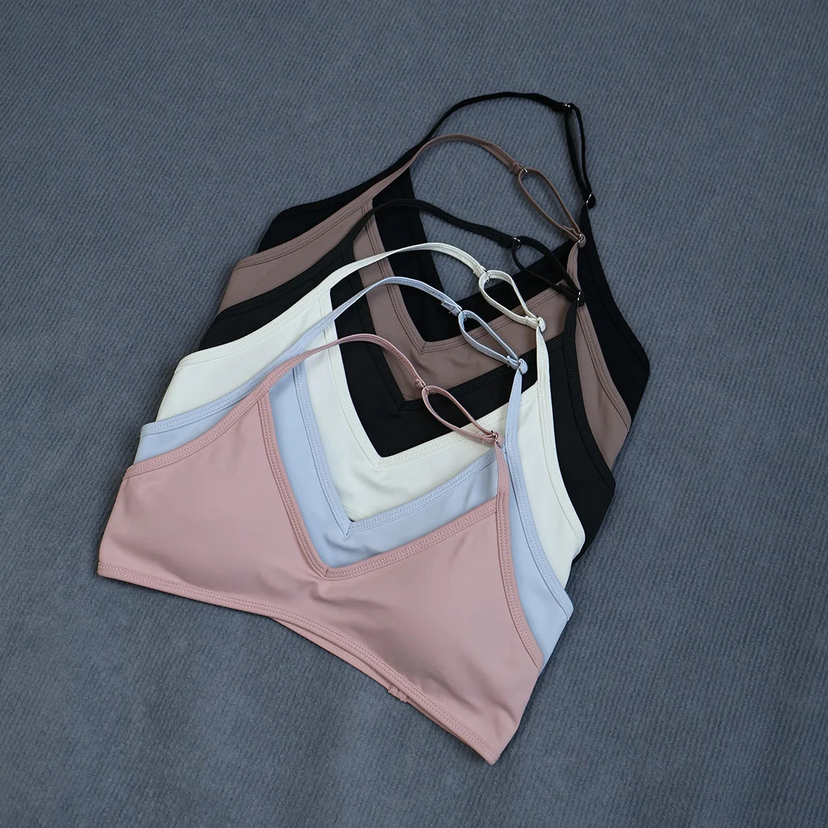 Four color variants of a seamless V-neck bikini top laid flat on a textured gray background. The colors shown are dusty pink, light blue, white, black, taupe, and brown.