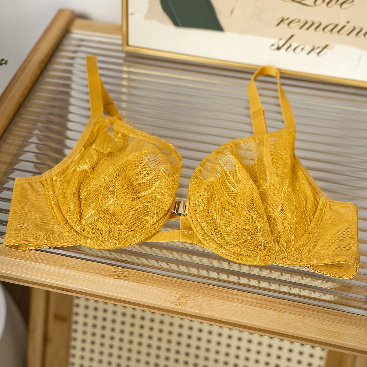 A yellow lace underwire bra with adjustable straps, displayed on a slatted wooden table with a framed print in the background.
