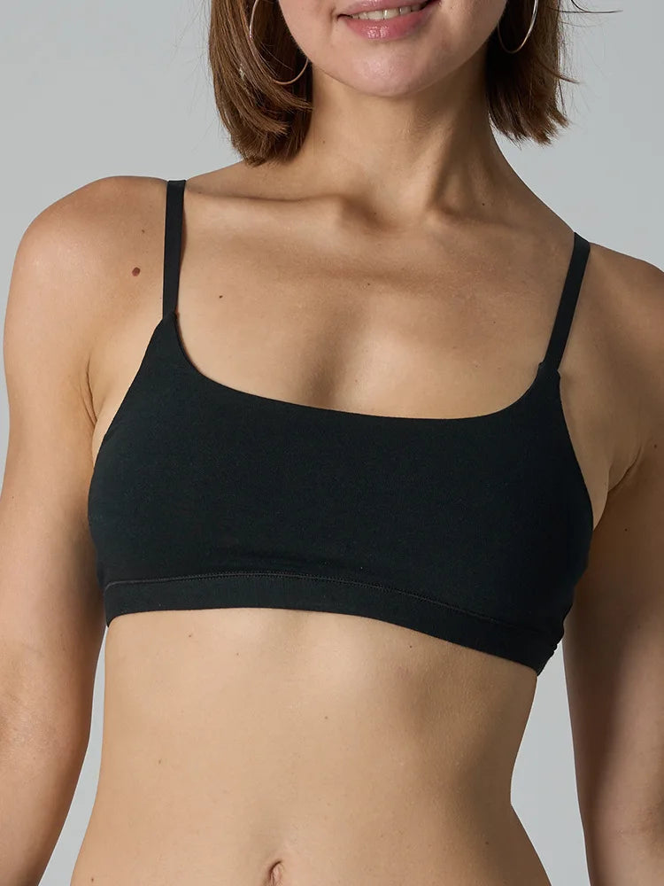 A close-up of a woman wearing a black bralette with thin straps and a wide, comfortable neckline against a plain light gray background.