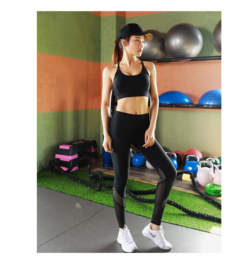A woman posing in a gym wearing a black sports bra and matching high-waisted leggings with sheer mesh panels on the knees, paired with white sneakers.