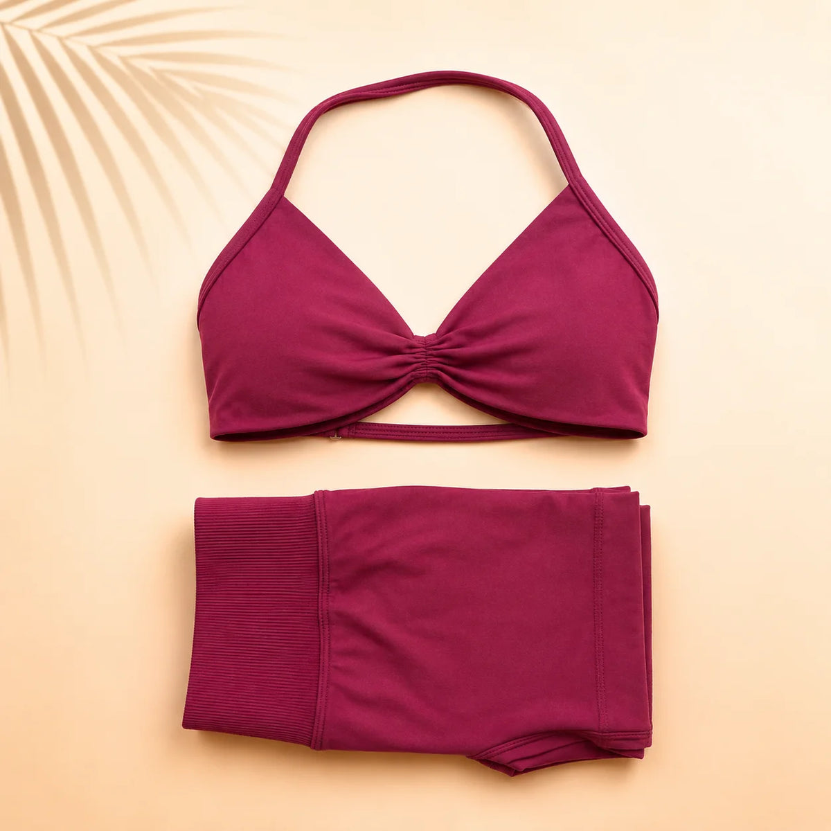 A two-piece bikini set in a deep magenta-purple color, featuring a halter-style triangle top with a twisted front detail and matching high-waisted shorts, laid flat on a warm beige background with a palm leaf shadow.