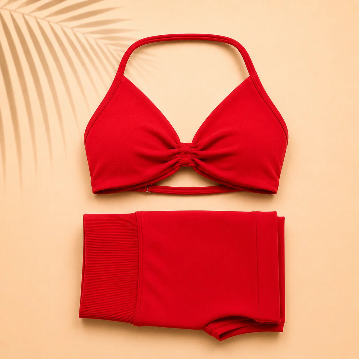 A bright red two-piece swimsuit set featuring a halter-style bikini top with a front twist detail and matching high-waisted bottoms, laid flat on a warm beige background with a palm leaf shadow.