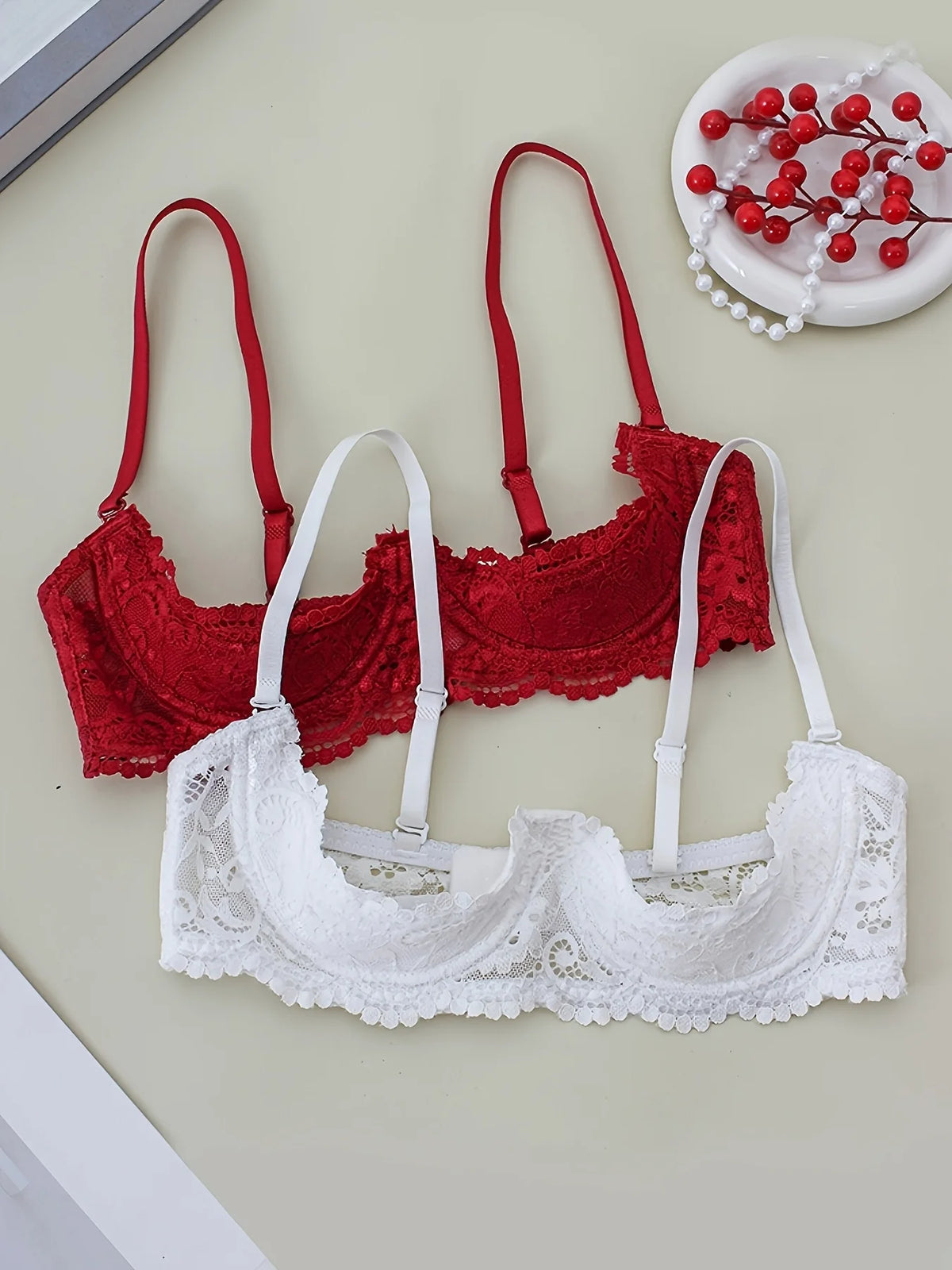 Two lace underwire bras laid flat on a light beige surface: one red bra at the back and one white bra in front. A small decorative plate with red berries and white pearls sits in the upper right corner.
