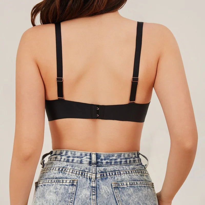 Back view of a woman wearing a black underwire bra with adjustable shoulder straps and a hook-and-eye closure, paired with light-wash denim jeans against a plain light beige background.