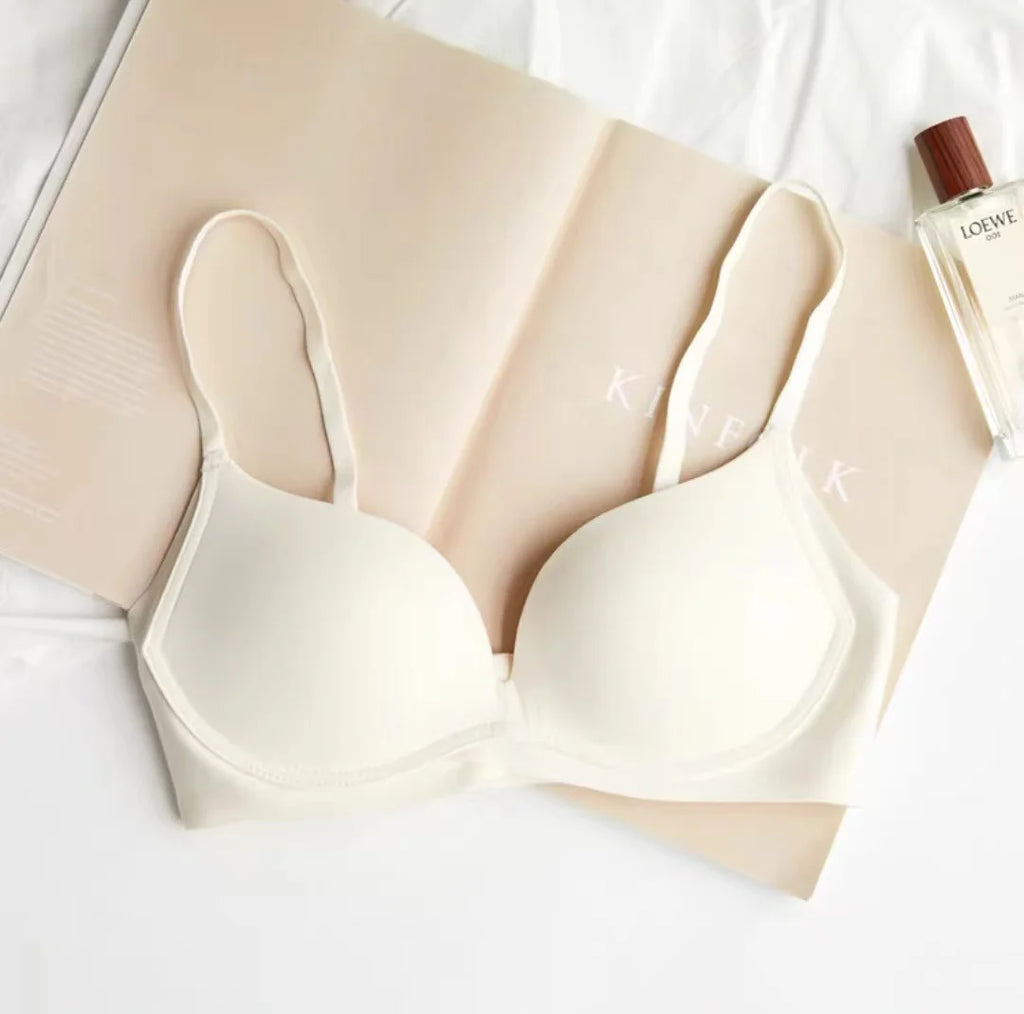 An off-white wireless bra laid flat on a beige book, with a perfume bottle at the top right and a white fabric surface underneath.