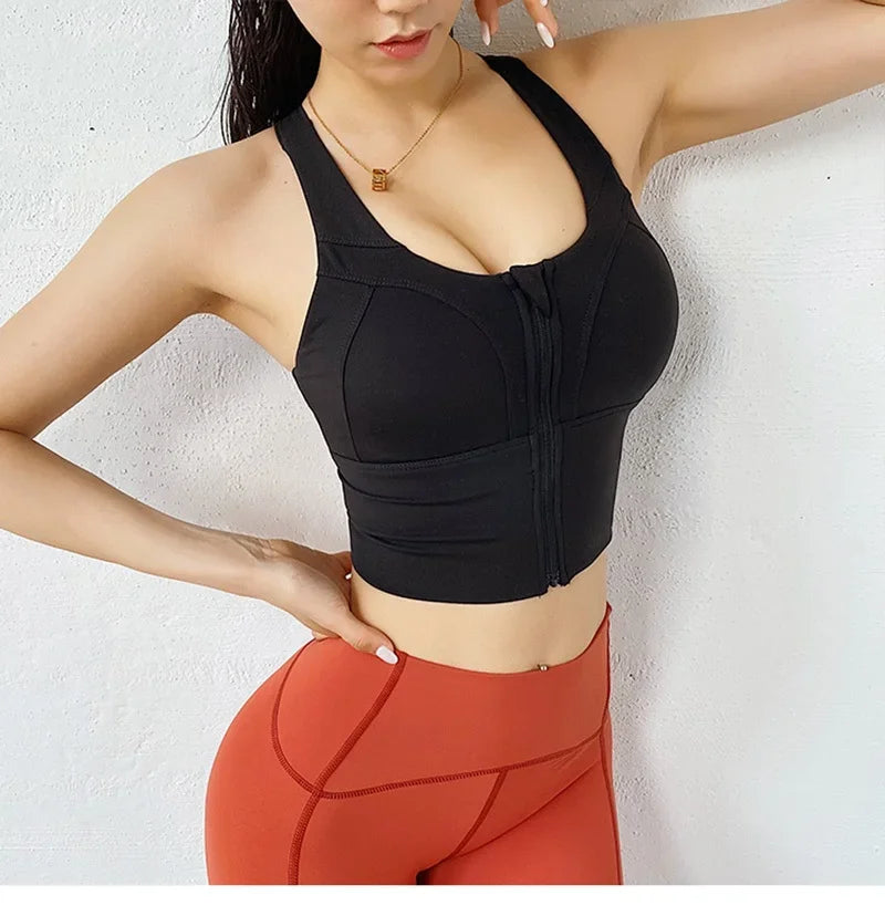 A woman wearing a black sports bra with a front zipper and underwire cups, paired with rust-orange high-waisted leggings, posing against a white textured wall.