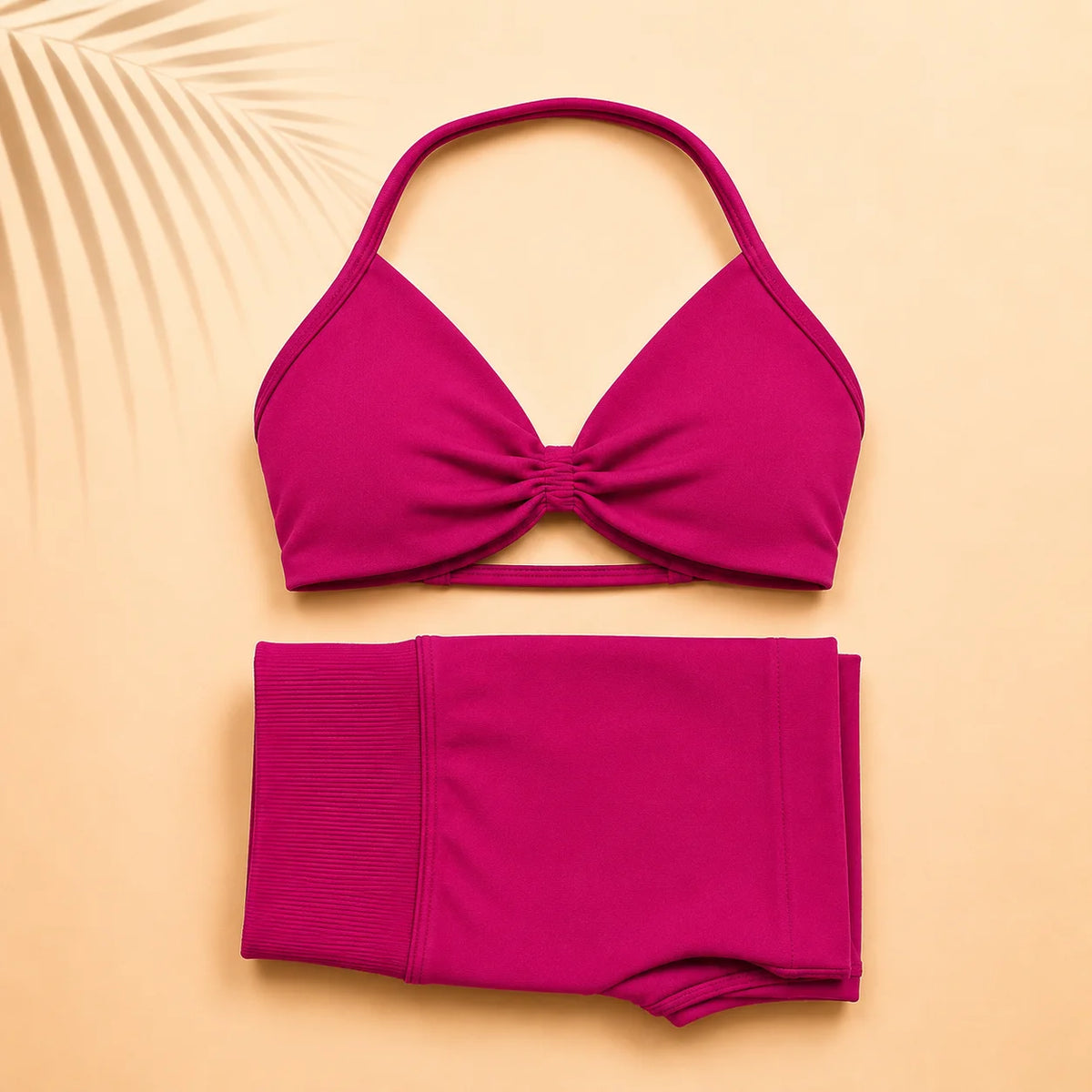 A two-piece bikini set in a vibrant magenta pink, featuring a halter-style triangle bikini top with a twisted front detail and matching high-waisted shorts, laid flat on a warm beige background with a palm leaf graphic in the corner.