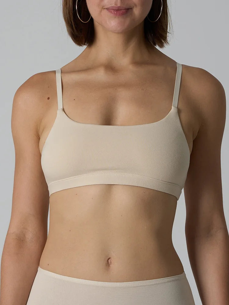 A woman wearing a beige seamless bralette with thin straps and a wide underband, shown from the chest to the hips against a light gray studio background.