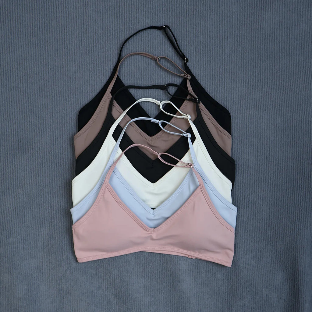 Five halter-style bralettes in soft neutral and pastel colors are stacked and laid flat on a textured gray background. The colors visible from front to back are dusty pink, light blue, white, black, and taupe.