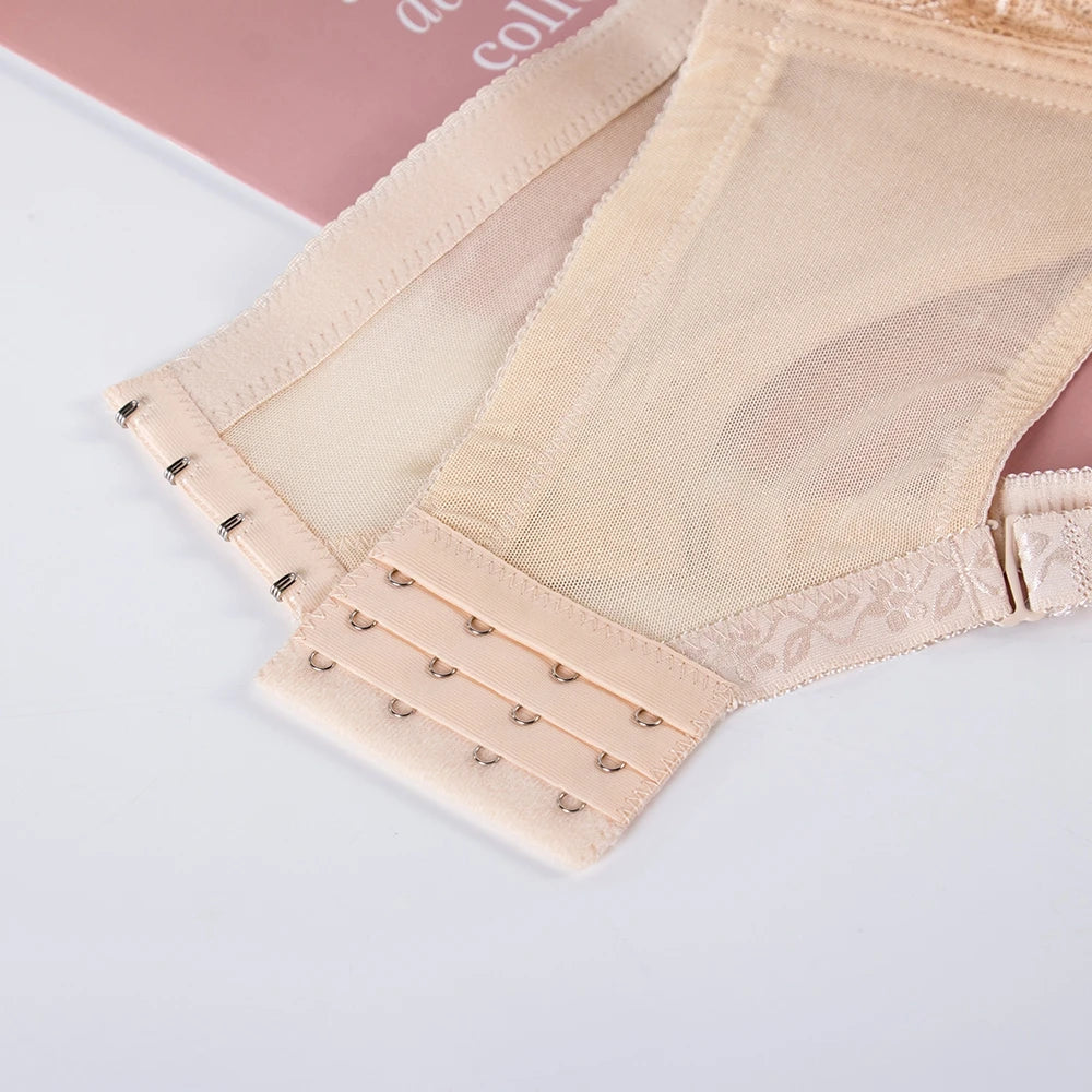 Close-up of a beige sheer mesh bra or panty with lace trim and adjustable hook-and-eye closure, laid flat on a white surface with a pink book corner visible in the background.