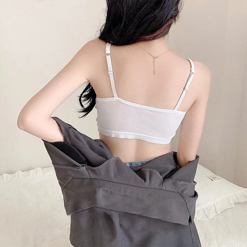A woman shown from behind wearing a white spaghetti-strap camisole bralette and holding a dark gray jacket over her shoulder in a softly lit indoor setting.