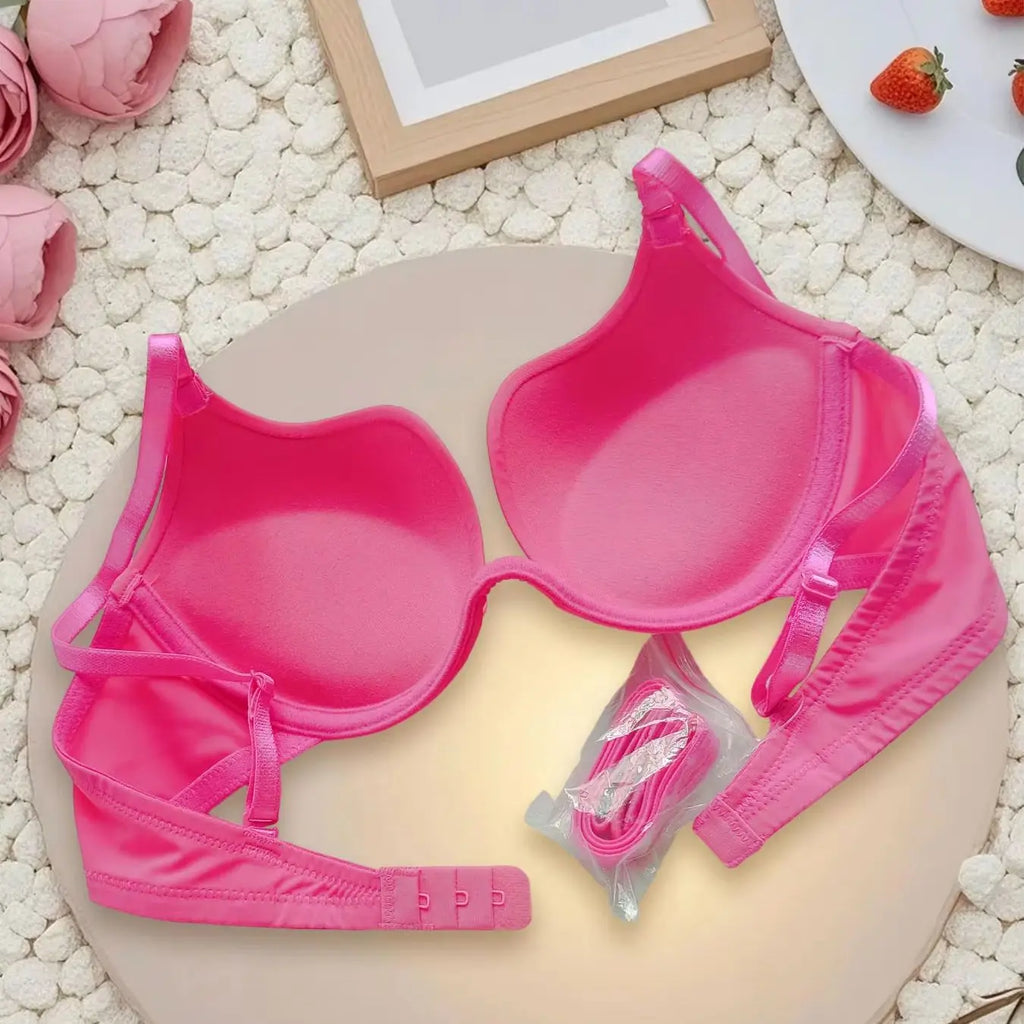 A bright pink molded-cup bra laid flat on a round beige board, with matching pink underwear pieces and a small packaged accessory nearby. The scene is styled with pink flowers, a wooden frame, and strawberries on a white textured surface.