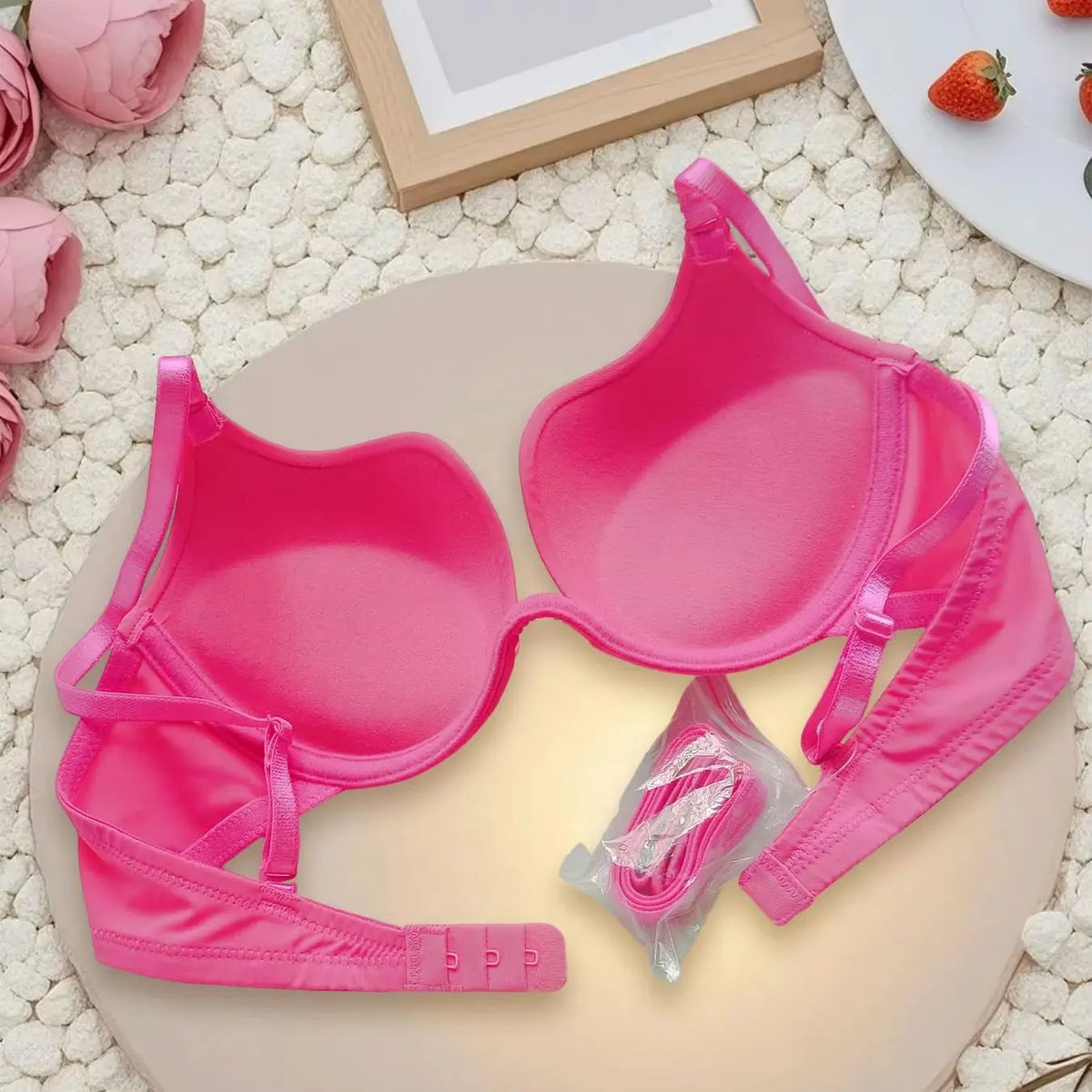 A bright pink molded-cup bra laid flat on a round beige board, with matching pink underwear pieces and a small packaged accessory nearby. The scene is styled with pink flowers, a wooden frame, and strawberries on a white textured surface.