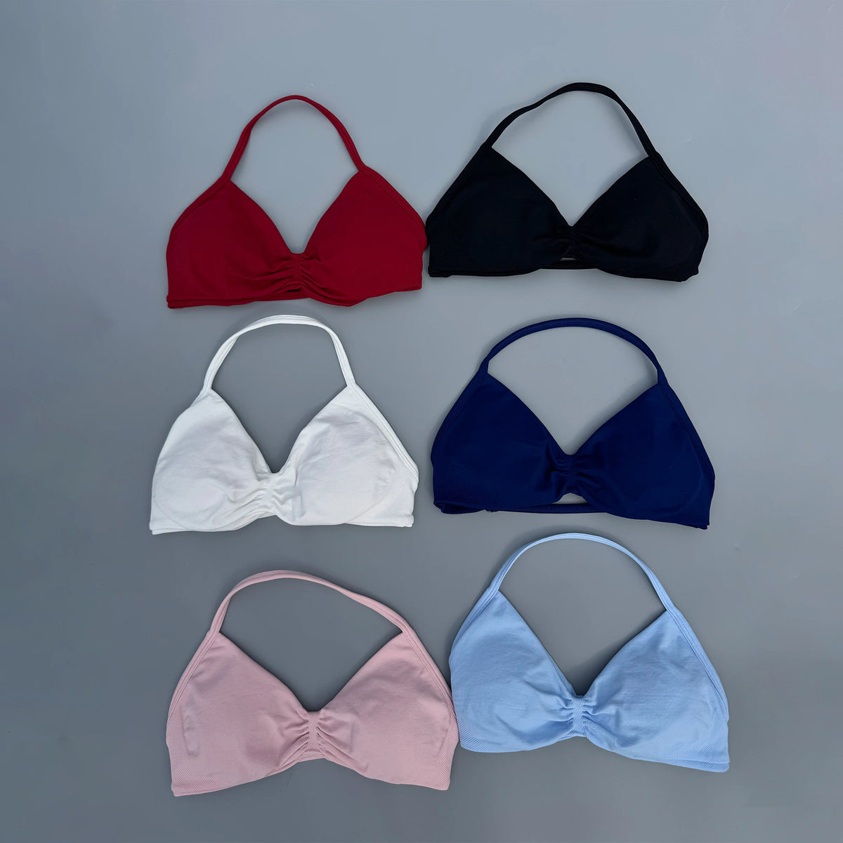 Six triangle bikini top cups arranged in a 2x3 grid on a smooth gray background, shown in red, black, white, navy, pink, and light blue. Each cup has a gathered front detail and thin straps.