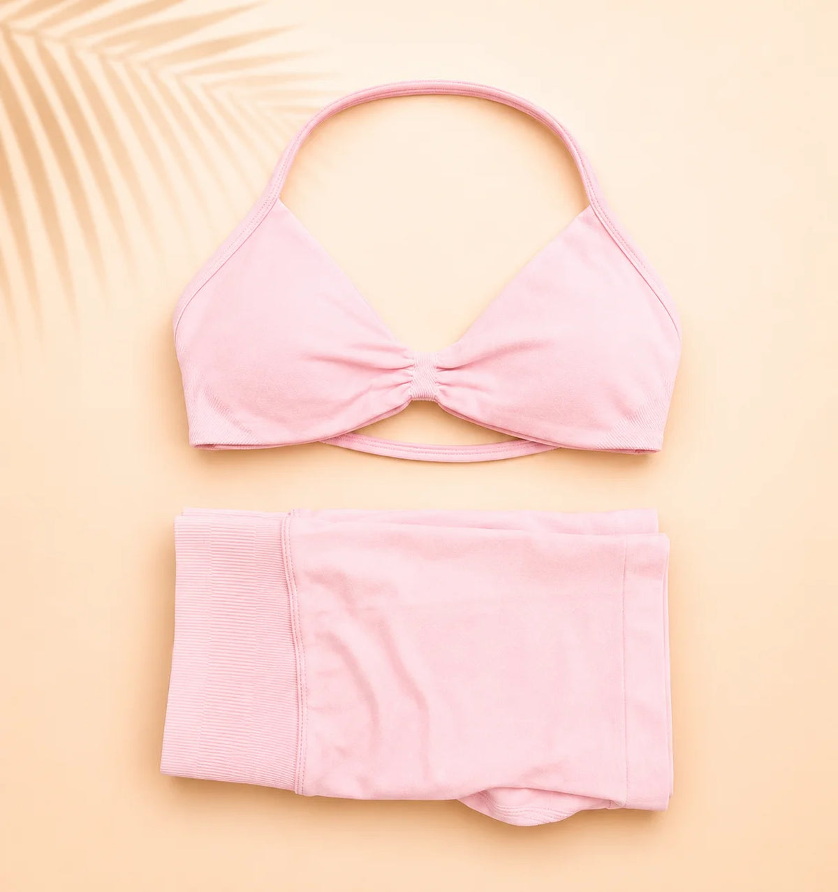 A light pink two-piece swimsuit laid flat on a warm beige background, featuring a halter-style triangle bikini top with a front twist detail and matching high-waisted bottoms.
