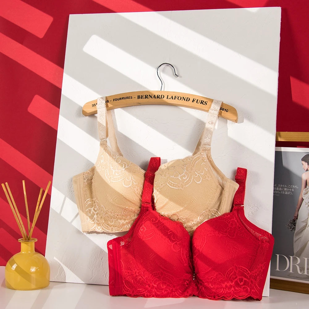 Two lace underwire bras displayed on a wooden hanger against a white board, with one beige bra on top and one red bra below. The setup includes a red wall backdrop, a yellow reed diffuser bottle, and a framed print on the side.