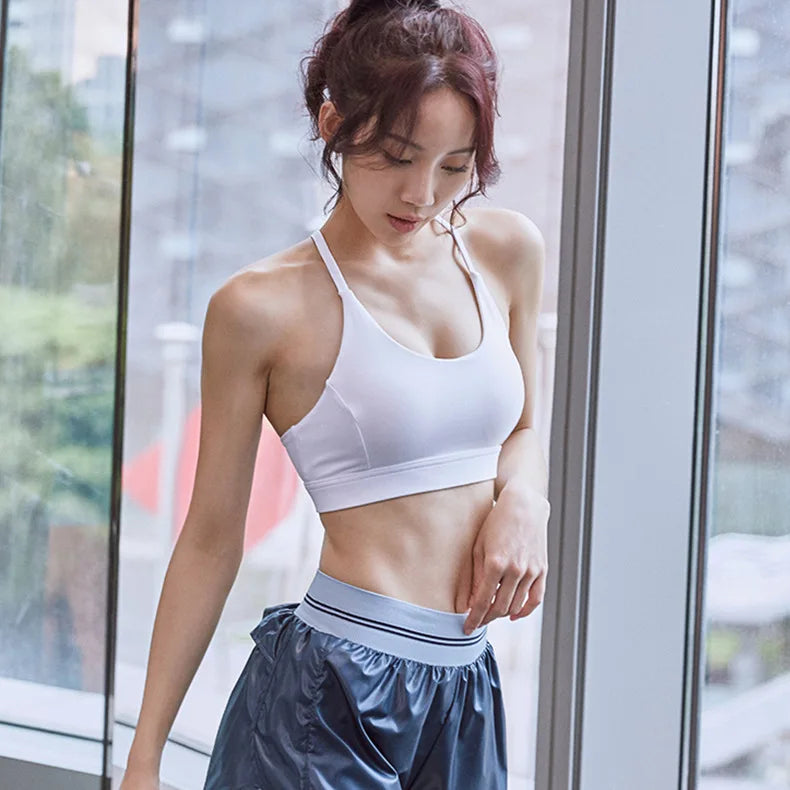 A woman wearing a white sports bra and shiny dark blue athletic shorts stands by a large window in a bright indoor setting.