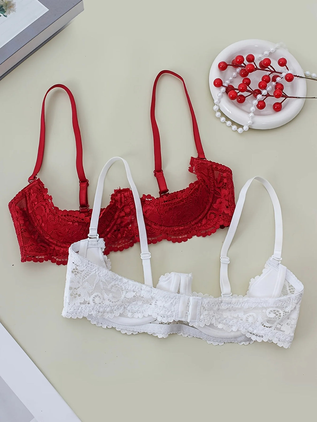 Two lace underwire bras, one red and one white, arranged on a light beige background with decorative props including a small dish of red berries and a book corner.