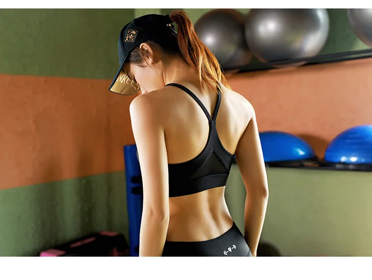 A woman in a gym wearing a black sports bra and matching black leggings, viewed from behind. She has a ponytail and wears a black baseball cap.