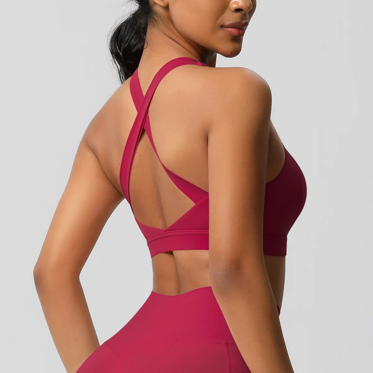 A woman shown from the back wearing a bright pink athletic sports bra with crisscross straps and a high-waisted matching bottom against a light gray studio background.