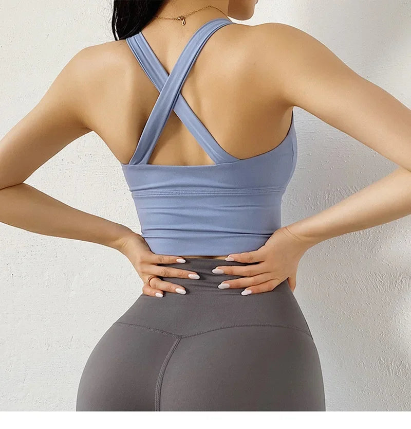 A woman shown from the back wearing a light blue athletic sports bra with crisscross straps and gray leggings, standing against a light textured wall.