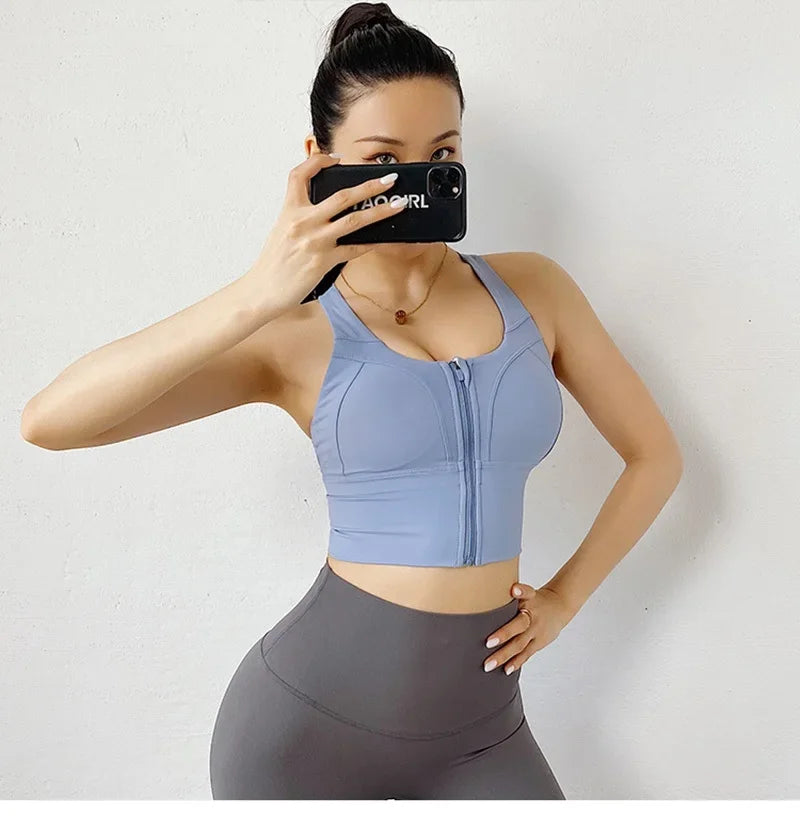 A woman takes a mirror selfie wearing a light blue athletic crop top with a front zipper and gray high-waisted leggings against a plain white wall.