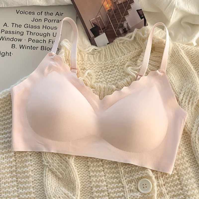 A pale blush pink wireless bra with scalloped edges and adjustable thin straps, laid flat on a cream knit cardigan with a magazine in the background.