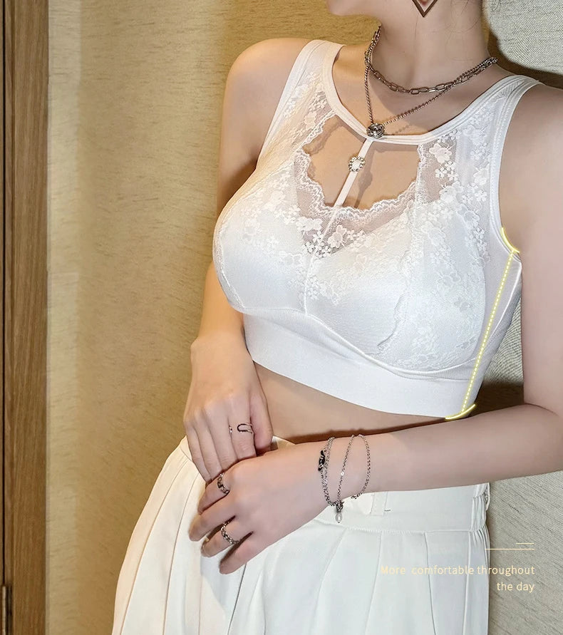 A woman wearing a white lace and mesh racerback crop top with underwire cups and a front keyhole detail, paired with a white skirt. She is wearing layered silver necklaces and bracelets, standing against a beige textured wall.