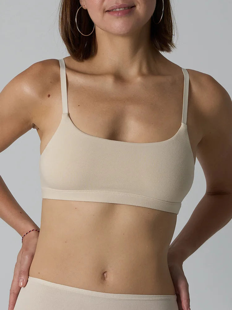 A woman wearing a beige wireless bralette with thin straps and a wide underband, shown from the shoulders to the hips against a light gray studio background.