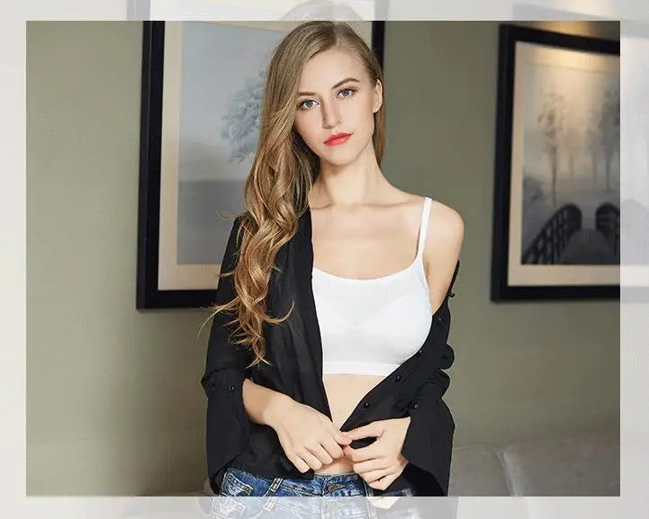 A woman with long wavy blonde hair is shown from the waist up, wearing a white ribbed camisole top, a black button-up shirt draped off her shoulders, and blue jeans. She is posed in front of a neutral-toned wall with two framed art prints.