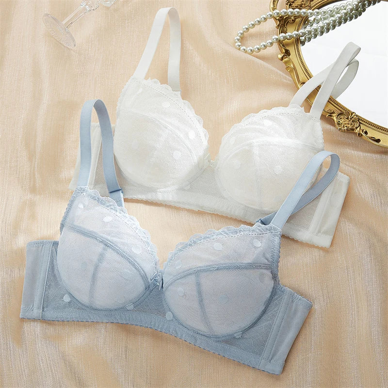 Two wireless lace-trimmed underwire bras laid flat on a beige satin-like fabric background. The front bra is light blue, and the bra behind it is white.