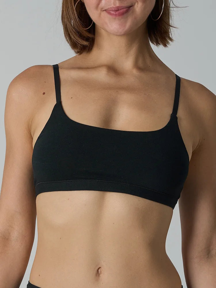 A close-up of a woman wearing a black scoop-neck bralette against a light gray studio background.