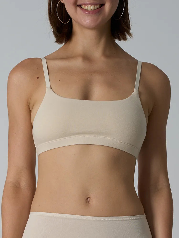 A woman wearing a beige seamless bralette with thin straps and a square neckline, paired with matching beige bottoms, against a solid gray background.