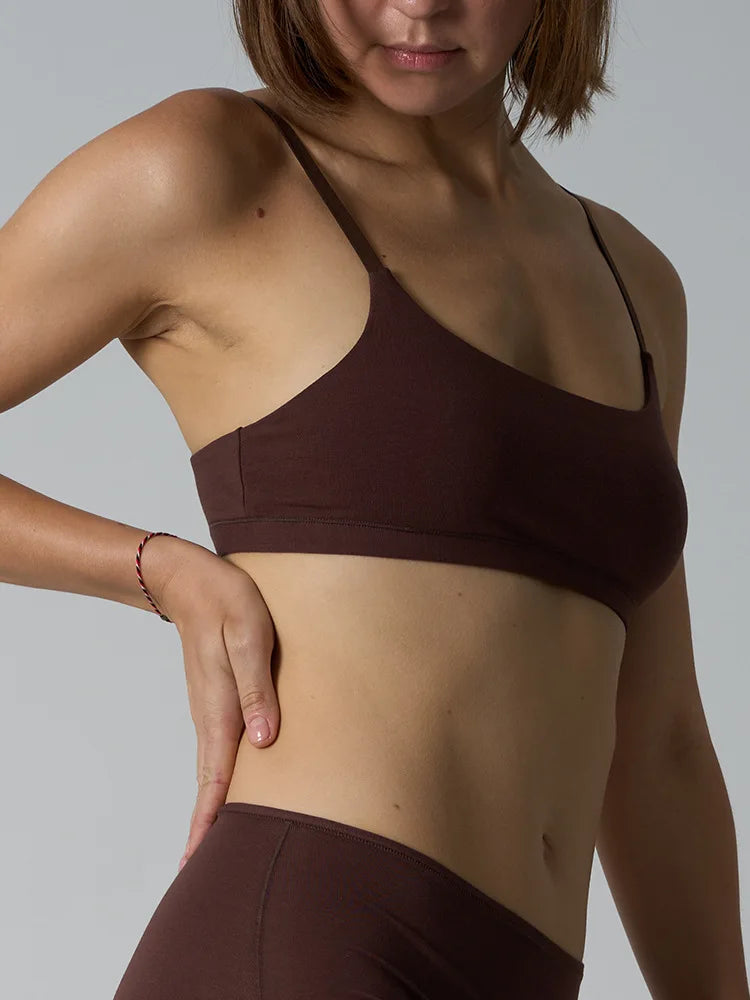 A close-up of a woman wearing a dark brown sports bra and matching high-waisted leggings against a solid gray studio background.