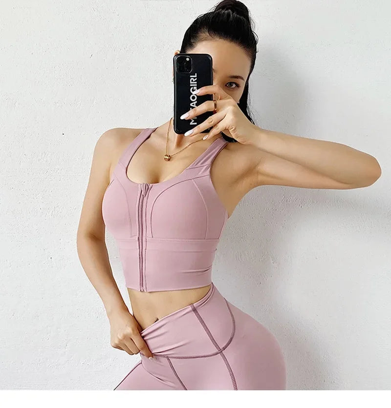 A woman takes a mirror selfie wearing a matching light pink activewear set consisting of a sports bra and high-waisted leggings. She is holding a black smartphone with 'YOCCURL' printed on the back, posing against a plain white wall.