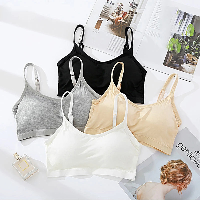 Four wireless bralettes in white, light gray, black, and beige, arranged in a flat-lay composition on a white surface with open magazines and a perfume bottle.
