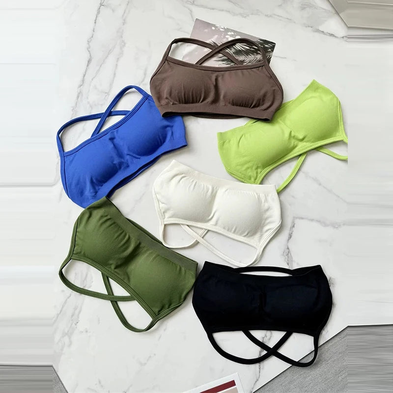 Six wireless sports bra or bikini top styles arranged on a light marble surface, shown in blue, brown, lime green, olive green, white, and black. Each top has a simple cup design with crossed straps.